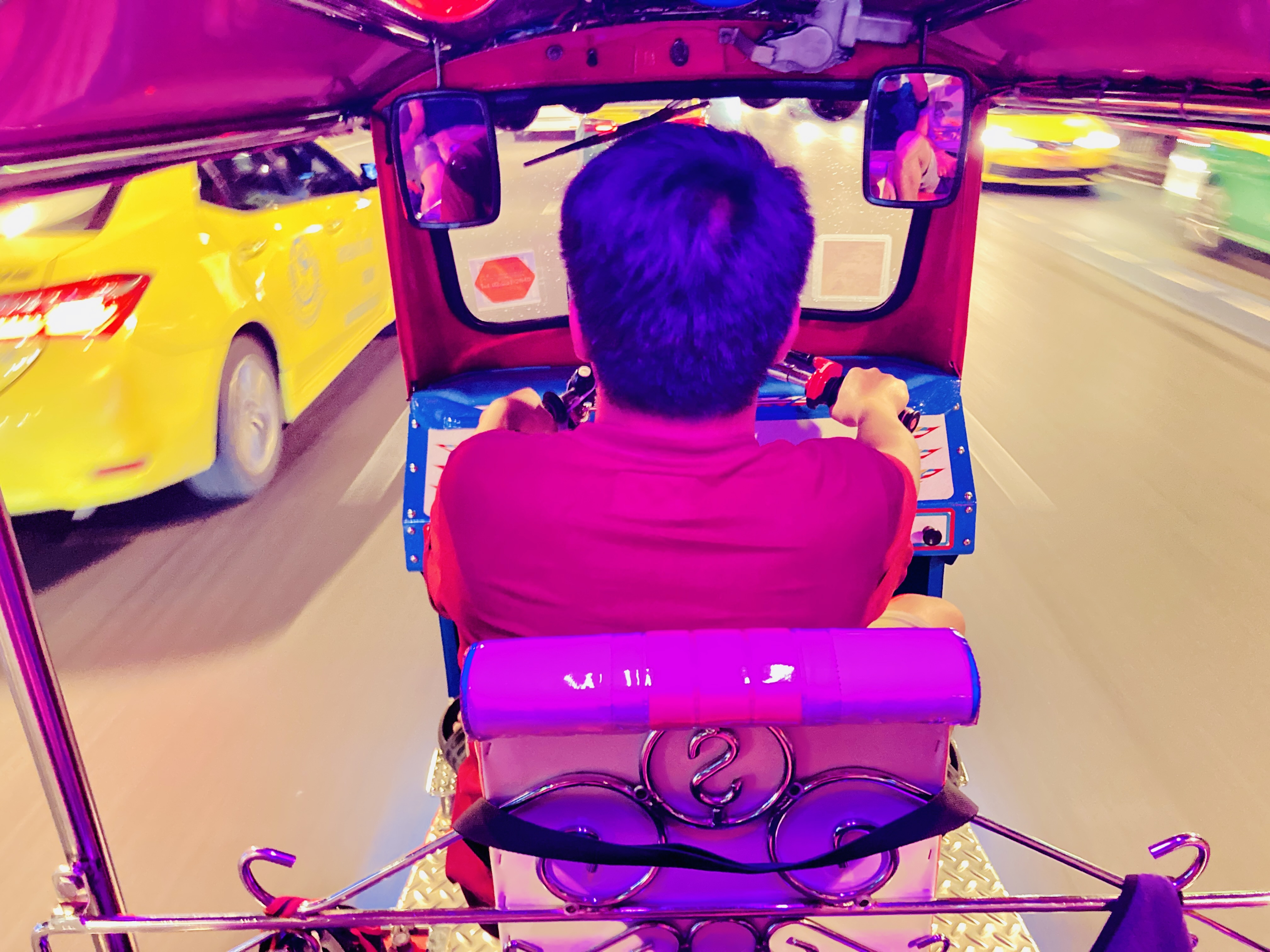interno di tuk-tuk di notte a Bangkok: autista in rosso, taxi giallo affianco, luci stradali sfocate per la velocità
