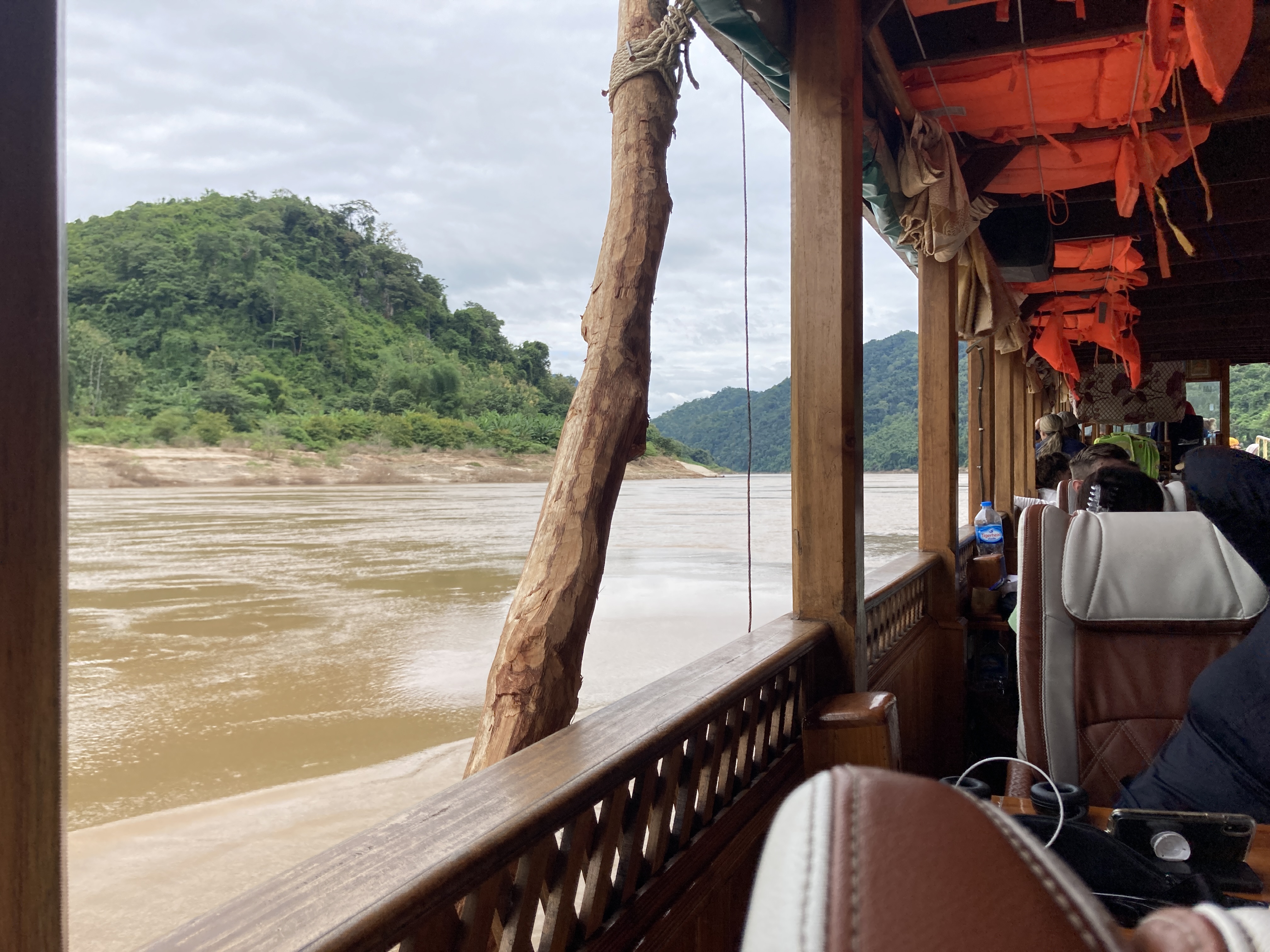 interno della slow boat sul Mekong: colonne di legno, giubbotti arancioni appesi, fiume torbido marrone, sponda verde boscosa, altri passeggeri