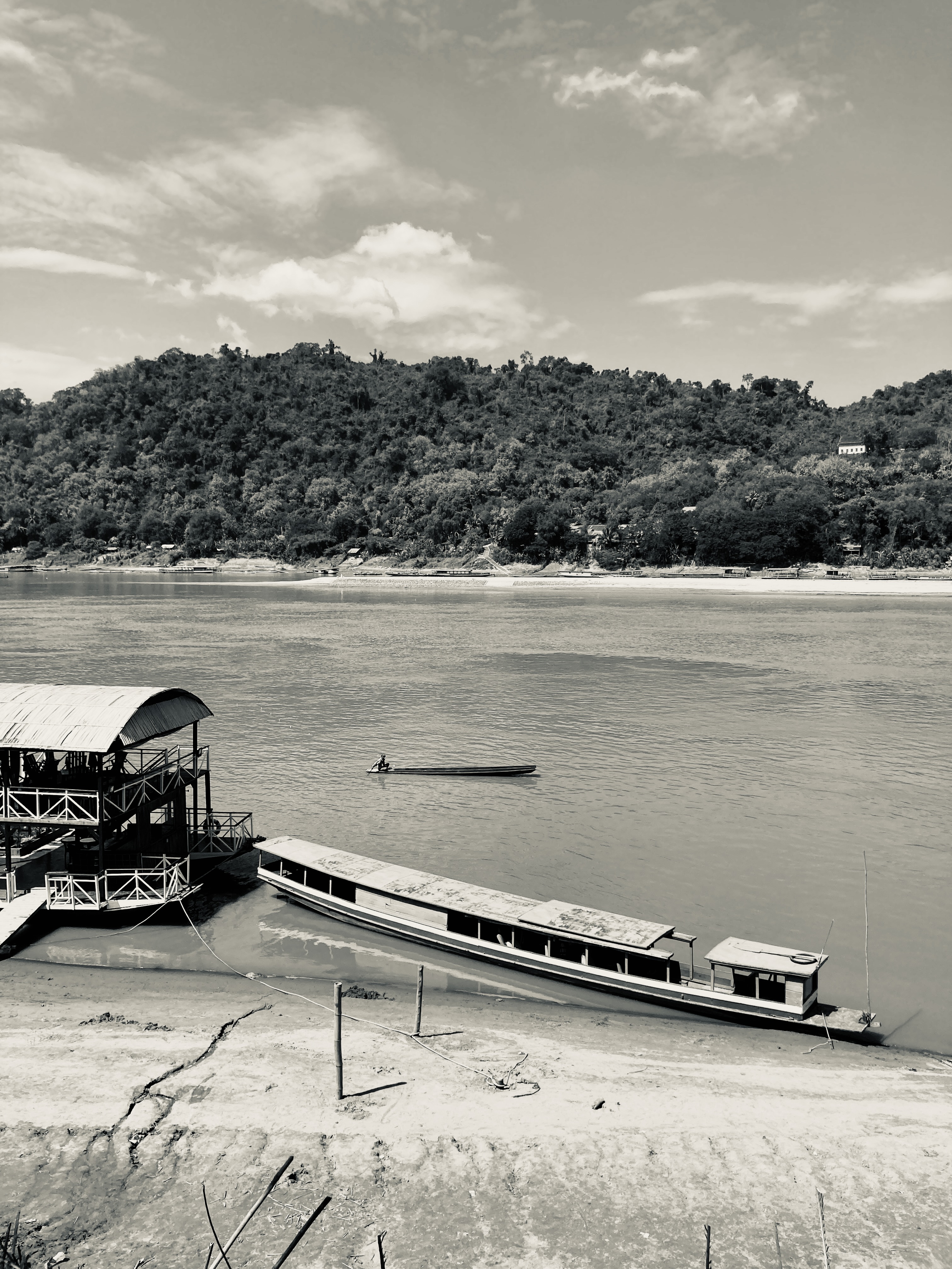 porto sul Mekong in bianco e nero: barca lunga ormeggiata sulla sabbia, struttura con tetto a sinistra, un rematore sull'acqua, colline boscose sullo sfondo