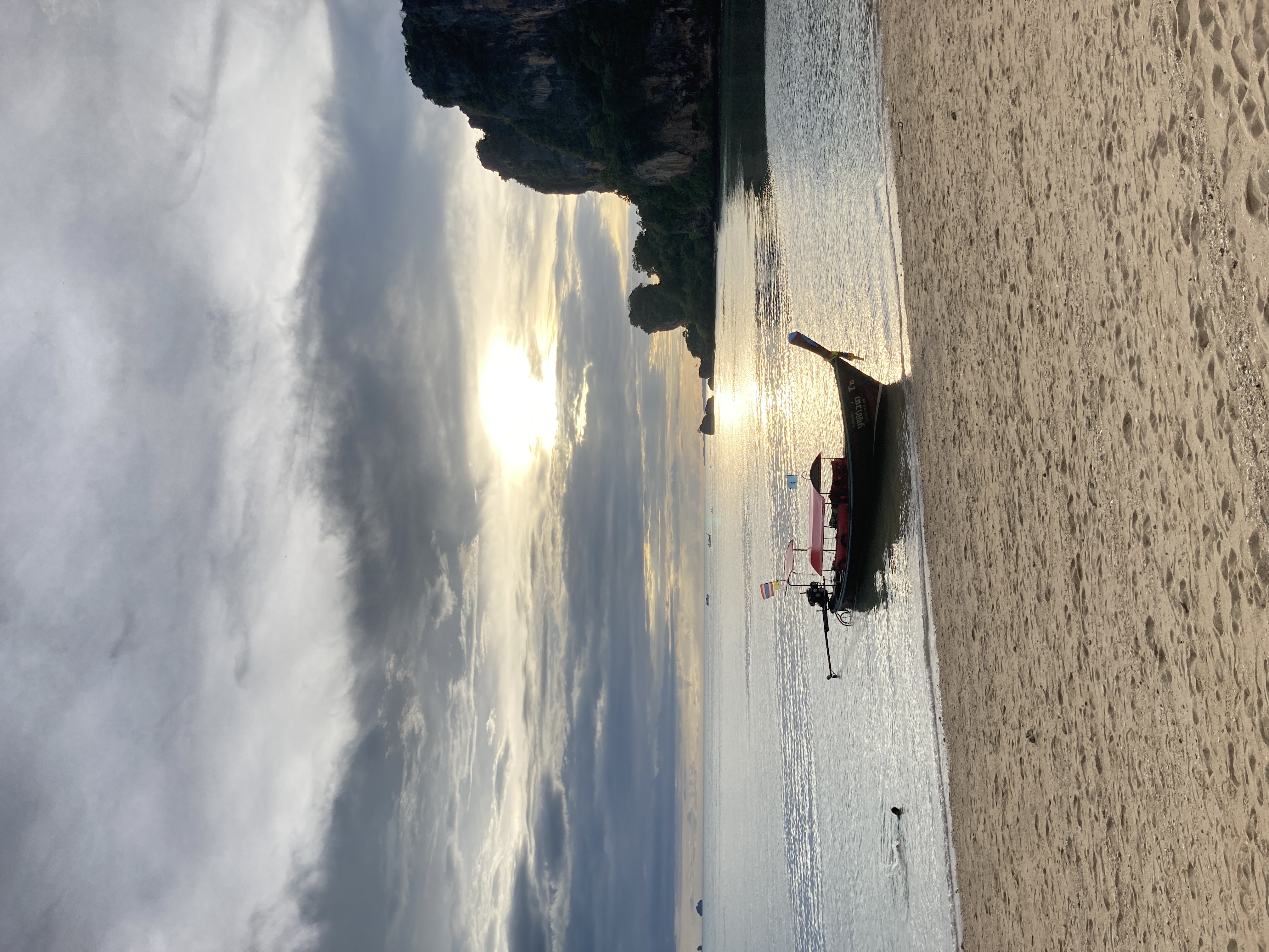 longtail boat ormeggiata sulla sabbia bianca di Railay, pareti calcaree scure sullo sfondo, sole che filtra tra le nuvole con riflesso dorato sul mare