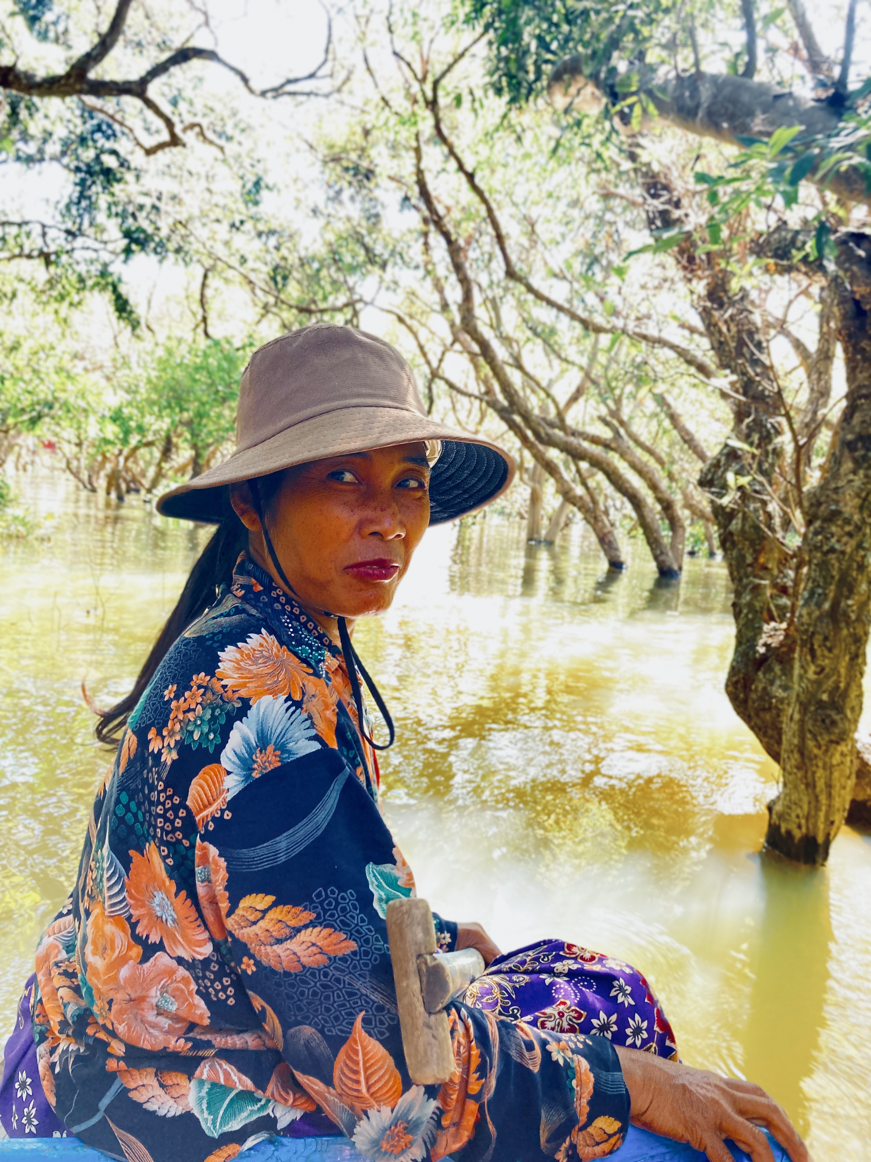 donna con cappello a tesa larga e vestito floreale blu-arancio su una barca, sullo sfondo alberi semi-sommersi nell'acqua giallo-verde del lago Tonle Sap
